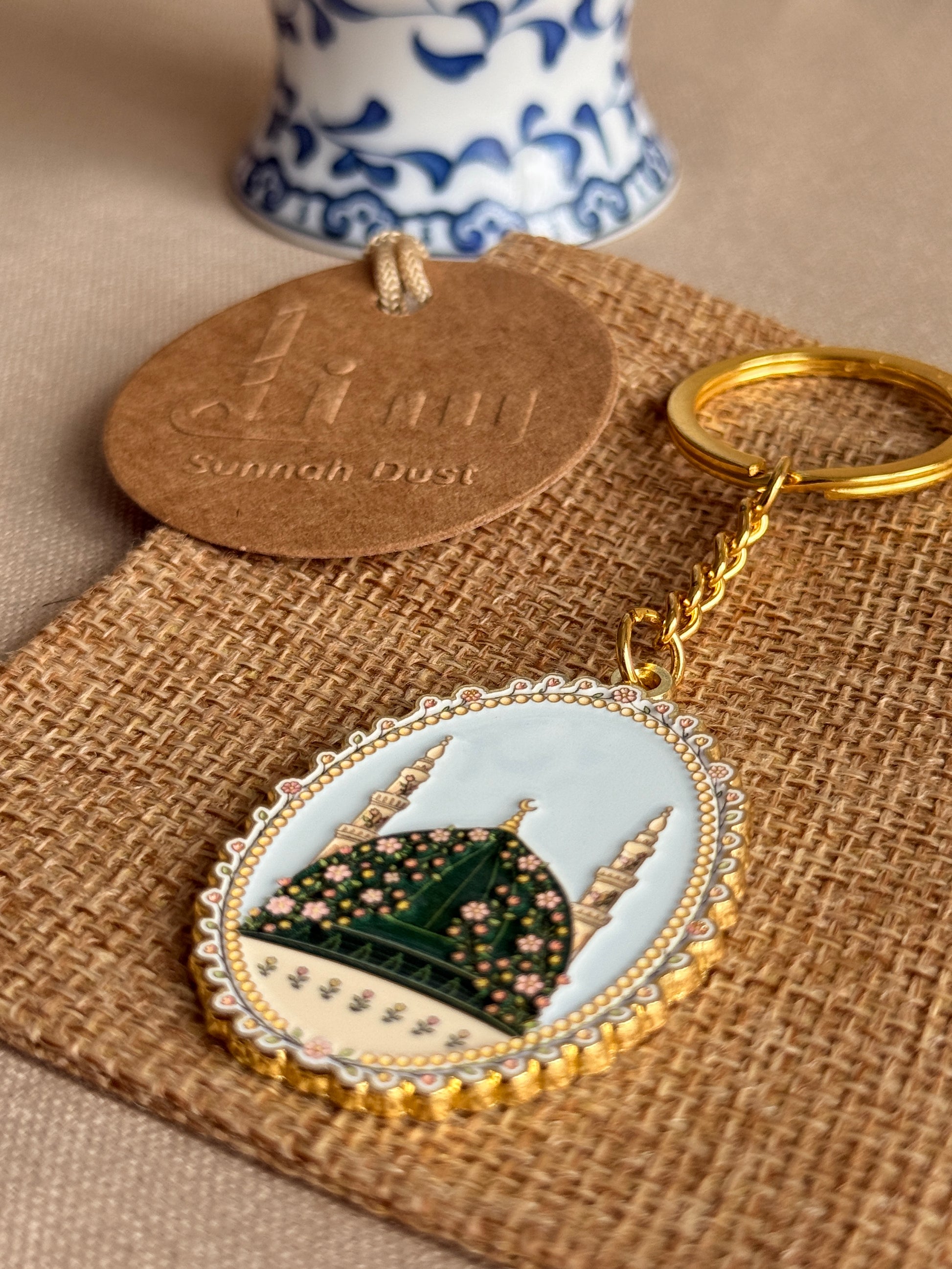 An oval enamel keychain with a gold-tone ring and chain, featuring an illustration of a green dome and minaret on a pale blue and cream background. The keychain is displayed on a burlap fabric next to its round cardboard tag, with a blue and white ceramic vessel blurred in the background.
