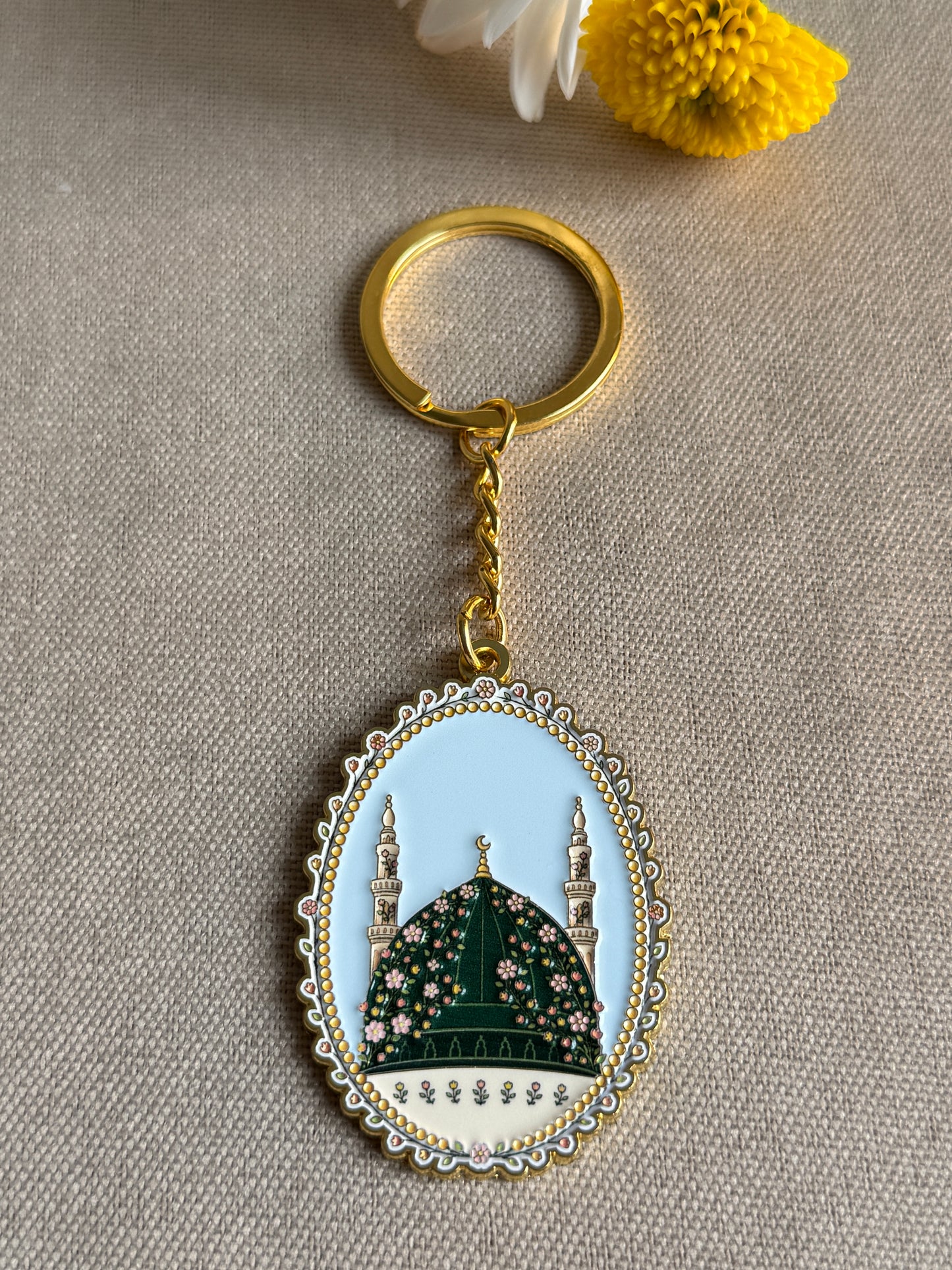 An oval keychain charm with a gold-tone ring and chain, featuring an illustration of a mosque dome and minarets on a light blue and cream background with floral detailing, displayed on a beige textured fabric surface.
