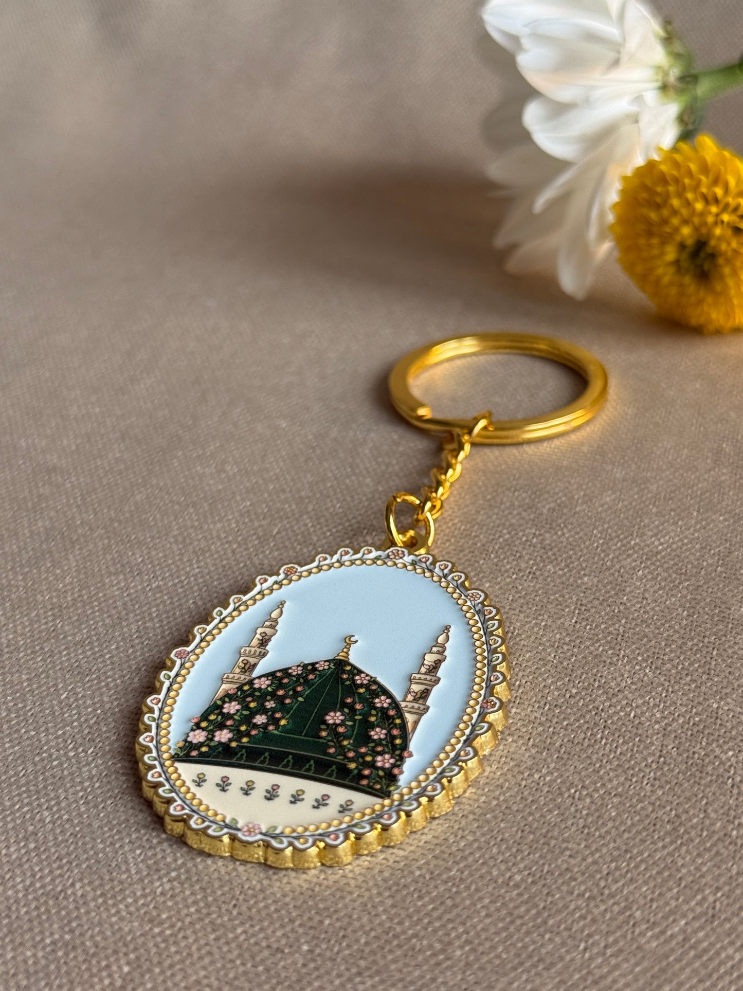 An oval enamel keychain featuring a mosque design with a light blue background, dark green dome with a light blue sky, and gold-toned metal border and key ring, photographed on a beige fabric surface with blurred flowers in the corner.